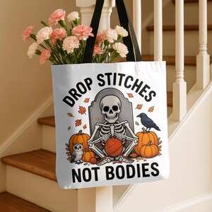 Op de afbeelding: Witte draagtas met zwarte hengsels. De tas heeft een skelet dat breit met een bol wol, pompoenen en de tekst "DROP STITCHES NOT BODIES". De tas is gevuld met roze en witte bloemen.