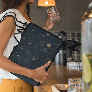 Op de afbeelding: Een donkerblauwe laptoptas met een gouden hemels ontwerp van zonnen, manen en sterren. De tas wordt vastgehouden door een persoon die een witte top met zwarte bies en een gele broek draagt. De achtergrond toont een bar met een glas water.