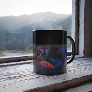 May include: A black heat-sensitive mug with a colorful mushroom and forest scene. The design features red and white spotted mushrooms, a blue forest, and a whimsical house. Steam rises from the mug, suggesting a hot beverage.