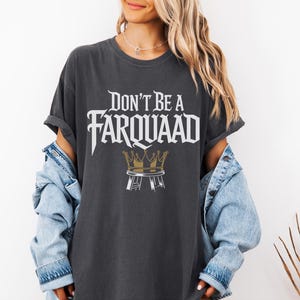 May include: Dark gray t-shirt with the text "Don't Be A Farquaad" in a stylized font. Below the text is a gold crown design. The shirt is worn with a light blue denim jacket.