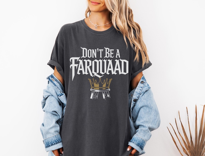May include: Dark gray t-shirt with the text "Don't Be A Farquaad" in a stylized font. Below the text is a gold crown design. The shirt is worn with a light blue denim jacket.