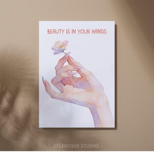 May include: A watercolor print featuring a hand holding a delicate flower. The text "BEAUTY IS IN YOUR HANDS" is displayed in red. The print has a soft, pastel color palette and is signed "SELENIQUE STUDIO".