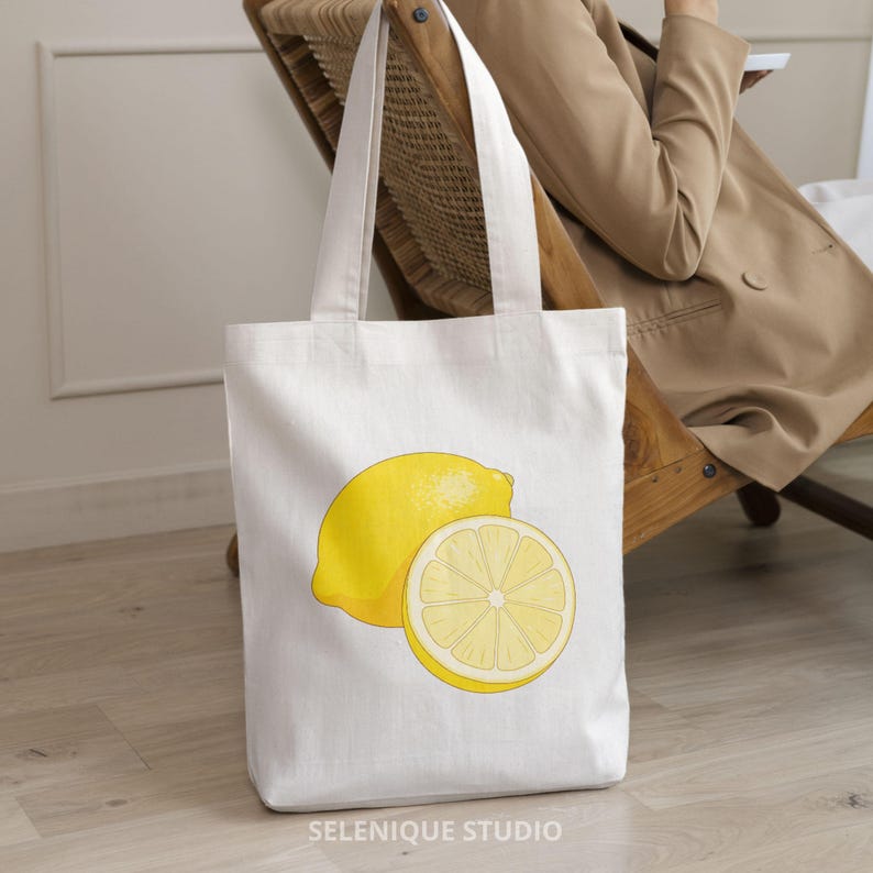 Puede incluir: Bolso tote blanco con una ilustraci&oacute;n de un lim&oacute;n amarillo y una rodaja de lim&oacute;n. El bolso tiene dos asas y est&aacute; hecho de tela clara. El texto "SELENIQUE STUDIO" est&aacute; impreso en la parte inferior del bolso.