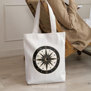 Pu&ograve; includere: Borsa tote bianca con un design a bussola nero e oro. La rosa dei venti &egrave; circondata da stelle e lune crescenti. La borsa ha manici lunghi e il testo "SELENIQUE STUDIO" in basso.