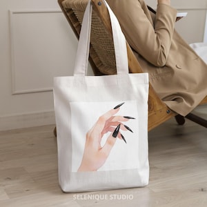 May include: White tote bag featuring a watercolor illustration of a hand with long, black fingernails. The bag has two long handles and the text "SELENIQUE STUDIO" at the bottom. The bag is made of a sturdy, off-white fabric.