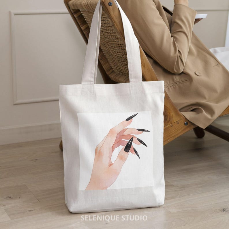 May include: White tote bag featuring a watercolor illustration of a hand with long, black fingernails. The bag has two long handles and the text "SELENIQUE STUDIO" at the bottom. The bag is made of a sturdy, off-white fabric.