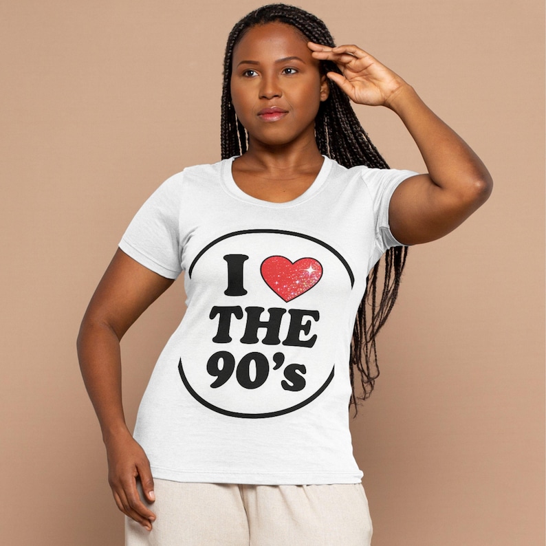Op de afbeelding: Wit T-shirt met een zwart ovaal ontwerp met de tekst "I ❤️ THE 90's". Het hart is rood en glinsterend. Het shirt heeft korte mouwen en een ronde hals.