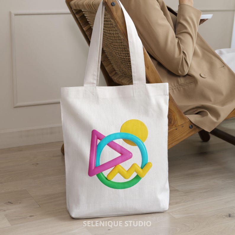 May include: White canvas tote bag with a colorful geometric design. The design includes a pink triangle, a blue circle, a yellow sun, and a green and yellow zig-zag shape. The bag has long handles and the text "SELENIQUE STUDIO" at the bottom.