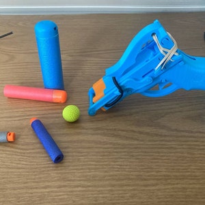 Puede incluir: Una pistola de juguete azul con una banda elástica y dardos azules, naranjas y grises. También hay una pequeña bola verde.