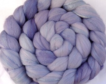 May include: A circular braid of soft, light purple wool roving. The wool is a blend of lavender and pale gray, creating a gradient effect. The braid is tightly woven, with each strand clearly visible. The texture appears fluffy and inviting.
