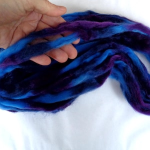 Op de afbeelding: Een streng handgeverfde merino wol in blauwe en paarse tinten. De wol wordt door een hand vastgehouden, waarbij de textuur en kleurvariaties worden benadrukt. Ideaal voor handwerkprojecten zoals spinnen of vilten.