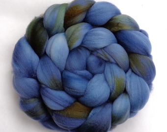 May include: A round, braided bundle of blue wool roving. The wool is a mix of shades, including deep blues, lighter blues, and hints of green and brown. The roving is tightly woven, creating a textured, tactile appearance.