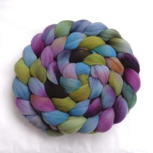 Può includere: Una spessa treccia circolare di lana cardata non filata caratterizzata da una tavolozza di colori variegati di viola intenso, magenta brillante, blu cielo, verde oliva e grigio scuro. La consistenza morbida e fibrosa è disposta a spirale.