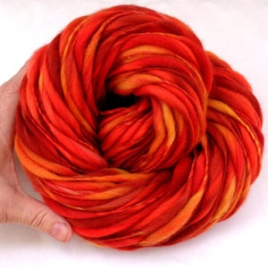 Puede incluir: Un ovillo de hilo teñido a mano en tonos de naranja, rojo y amarillo. El hilo está dispuesto en un círculo suelto, mostrando las variaciones de color.