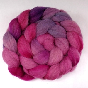Op de afbeelding: Een gevlochten bundel roze en paarse wolroving. De wol is een mix van tinten, met een verloop van donkerpaars naar lichtroze.