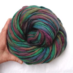 Könnte beinhalten: Ein großer Knäuel farbenfroher Roving-Wolle in Türkis-, Grün-, Lila- und Brauntönen. Die Wolle wird in einer Hand gehalten und zeigt ihre weiche Textur und die lebendigen Farbvariationen. Ideal für Kunstprojekte mit Fasern.