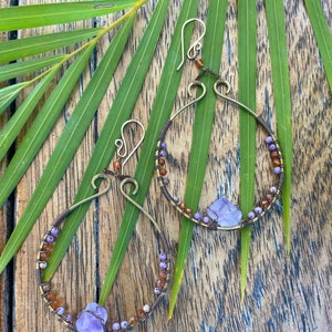 May include: A pair of handmade earrings with a teardrop shape. The earrings are made of brass wire and feature amethyst stones and brown and purple beads.