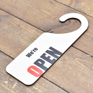 May include: White wooden door hanger with a hook at the top. The hanger has black text that says "We're OPEN" with the "O" in red.