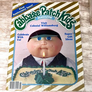 May include: A Winter 1986 Cabbage Patch Kids Magazine featuring a doll with blue eyes and a dark cap behind a yellow cake with a single lit candle. The cover text includes First Anniversary Issue, Visit Colonial Williamsburg, and Super Gift Ideas.