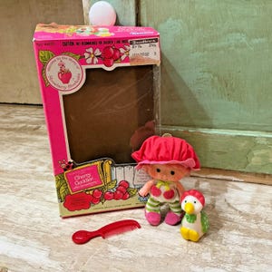 May include: A vintage Strawberry Shortcake Cherry Guddler doll set. The set includes a doll with a red hat, a small penguin figure, a red comb, and a box with the text "Cherry Guddler with Goodberry". The box is pink and has a clear window.