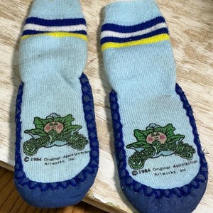 May include: A pair of light blue slipper socks with dark blue trim and a dark blue, white, and yellow striped cuff. Each sock features a Cabbage Patch Kids logo with the text 1984 Original Appalachian Artworks, Inc. printed on the sole.