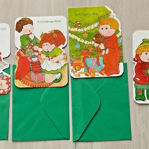 May include: Four rectangular Christmas greeting cards with rounded corners, each featuring illustrations of children with yarn hair. The cards display the text To A Sweet Girl, A Christmas Wish, To A Special Boy, and It's Christmas, paired with three green envelopes.