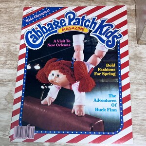 May include: A 1987 Cabbage Patch Kids magazine featuring a doll performing a handstand on a balance beam. The cover has red and white diagonal stripes, blue borders with white stars, and text including Kids Olympics, A Visit To New Orleans, Bold Fashions For Spring, and The Adventures Of Huck Finn.