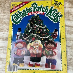 May include: A Winter 1985 Cabbage Patch Kids Magazine premier issue with a yellow border. The cover features three dolls in winter clothing holding books in front of a decorated Christmas tree, a snowman, and a nutcracker. Text includes "FREE GIFT INSIDE".
