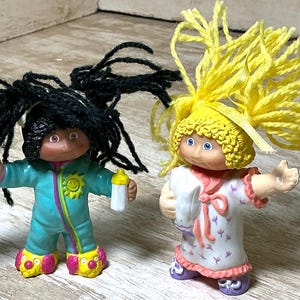 May include: Two small plastic dolls with yarn hair. The left doll has black hair, a teal jumpsuit with a yellow sun emblem, and holds a white bottle. The right doll has yellow hair, a white nightgown with purple floral patterns, and holds a white pillow.