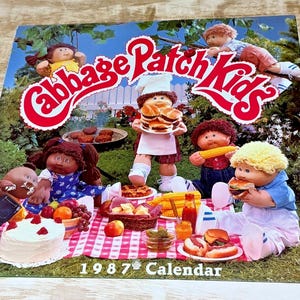 May include: A 1987 Cabbage Patch Kids calendar featuring a group of dolls having a picnic on a red and white checkered blanket. The dolls are surrounded by various food items including a cake, burgers, corn, and fruit in a garden setting.