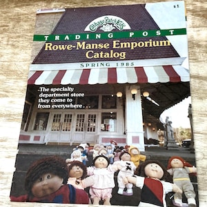 May include: A Cabbage Patch Kids Trading Post Rowe-Manse Emporium Catalog from Spring 1985. The cover features a storefront with a red and white striped awning and a group of various Cabbage Patch Kids dolls standing in front of the building entrance.
