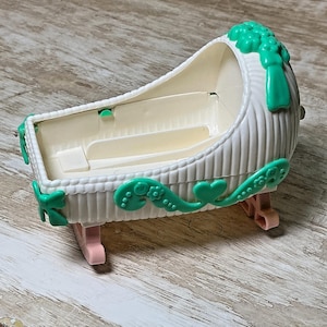 May include: A white plastic toy cradle with a ribbed exterior texture. The cradle features bright green decorative accents including floral and heart-shaped molded details along the sides and top. It is supported by two light pink rocking legs.