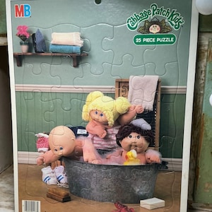 May include: A 25-piece Cabbage Patch Kids jigsaw puzzle featuring two dolls in a metal wash tub with a yellow rubber duck. The scene includes a shelf with an iron and towels, a washboard, and the text MB, Cabbage Patch Kids, 25 Piece Puzzle, and Ages 3 to 7.