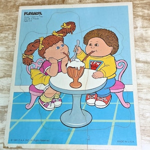 May include: A 1991 Playskool Sweet Treats for Two jigsaw puzzle featuring two Cabbage Patch Kids characters sitting at a round table sharing a sundae. The puzzle includes nine pieces and depicts the characters with brown hair, colorful clothing, and sneakers.