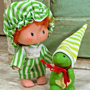 May include: A small doll with bright orange hair wearing a green and white striped bonnet and matching dress, standing next to a small green turtle figurine wearing a green and white striped pointed hat and a thin red ribbon tied around its neck.