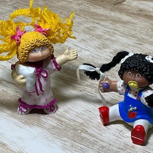 May include: Two small plastic figurines. One doll has bright yellow yarn hair, blue eyes, and wears a white nightgown with pink accents while holding a white pillow. The other doll has black yarn pigtails, a yellow pacifier, and a blue dress.