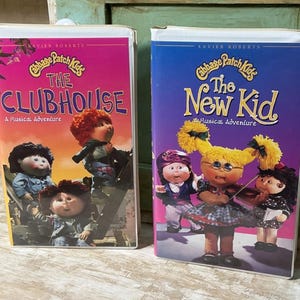 May include: Two Cabbage Patch Kids VHS tapes titled The Clubhouse: A Musical Adventure and The New Kid: A Musical Adventure. The first cover is pink with three dolls, while the second is purple featuring three dolls, one playing a violin.
