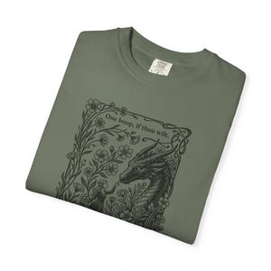 May include: An olive green t-shirt featuring a black ink-style graphic of a dragon and a person surrounded by flowers. The text above the dragon reads, One boop, if thou wilt. The shirt has a crew neckline and a visible size L tag at the collar.