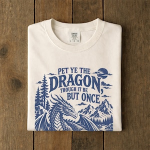 May include: A cream-colored t-shirt featuring a blue graphic print of a dragon, mountains, and pine trees. The text reads, "PET YE THE DRAGON, THOUGH IT BE BUT ONCE." The shirt has a crew neckline and a Comfort Colors brand label at the inner collar.