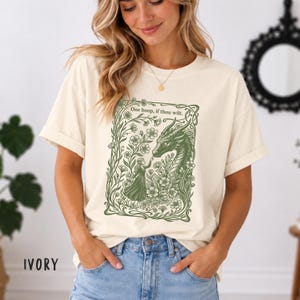 May include: An ivory short-sleeve t-shirt featuring a green graphic print of a person reaching toward a dragon surrounded by flowers and vines. The text "One boop, if thou wilt." is printed in green above the illustration.
