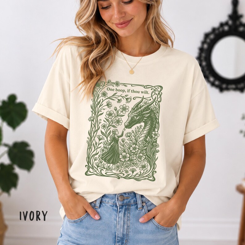 May include: An ivory short-sleeve t-shirt featuring a green graphic print of a person reaching toward a dragon surrounded by flowers and vines. The text "One boop, if thou wilt." is printed in green above the illustration.