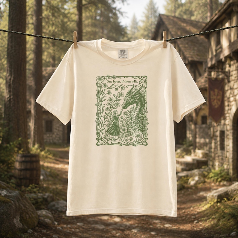 May include: A cream-colored short-sleeve t-shirt featuring a green graphic print of a person reaching toward a dragon surrounded by flowers. The text "One boop, if thou wilt." is printed above the illustration within a floral border.