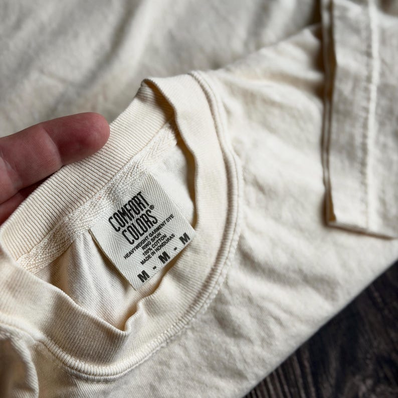 May include: A cream-colored t-shirt with a ribbed crew neckline. A white fabric label is sewn into the inner collar, featuring the black text Comfort Colors, Heavyweight Garment Dye, Ring Spun, 100% Cotton, Made in Honduras, and size M.