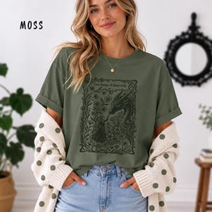 May include: A moss green t-shirt featuring a black graphic print of a person touching a dragon's nose surrounded by flowers and vines. The text "One boop, if thou wilt." is printed above the dragon. The shirt has short sleeves and a crew neckline.