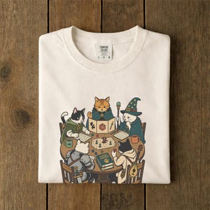 May include: An off-white t-shirt featuring a graphic of four cats playing a tabletop role-playing game around a round wooden table. The cats are dressed in fantasy attire, including a wizard hat and armor, with a book labeled Player's Handbook.