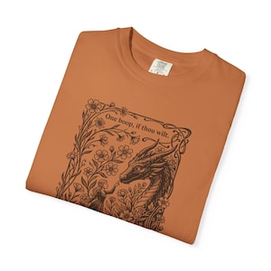 May include: A burnt orange t-shirt featuring a dark brown graphic print of a dragon and a person surrounded by flowers. The text "One boop, if thou wilt." is printed above the dragon. The shirt has a crew neckline and a size L label.