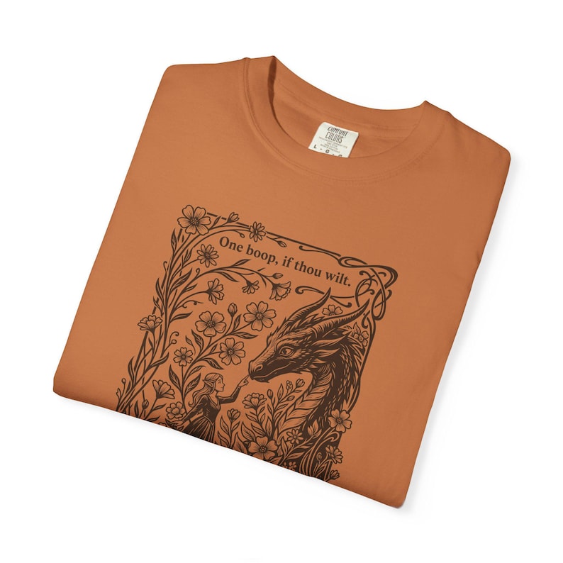 May include: A burnt orange t-shirt featuring a dark brown graphic print of a dragon and a person surrounded by flowers. The text "One boop, if thou wilt." is printed above the dragon. The shirt has a crew neckline and a size L label.