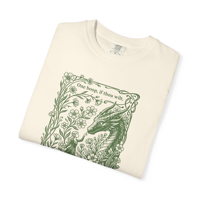 May include: An off-white t-shirt featuring a dark green graphic print of a person touching a dragon's nose. The design is framed by flowers and vines, with the text "One boop, if thou wilt." printed at the top. The shirt has a crew neckline.