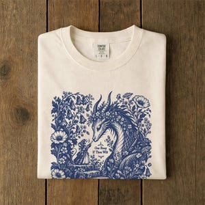 May include: An off-white t-shirt featuring a blue graphic of a small mouse reaching toward a dragon surrounded by flowers. The text "One Boop If Thou Wilt" is printed below the mouse. The shirt has a Comfort Colors brand neck label.