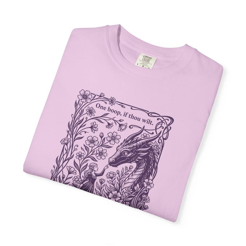 May include: A light pink t-shirt featuring a dark purple graphic print of a dragon and a person touching noses within a floral frame. The text "One boop, if thou wilt." is printed above the dragon. The shirt has a crew neck and a label.